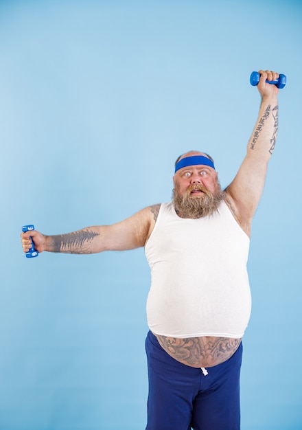 Photo shocked mature obese man with beard does exercises with dumbbells training hard on light blue background in studio