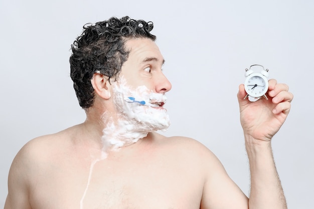 Shocked middle-aged white man with toothbrush in his mouth, shave, washing head, looks in profile on alarm clock in hand and hurry, being late for work or meeting, overslept on white background