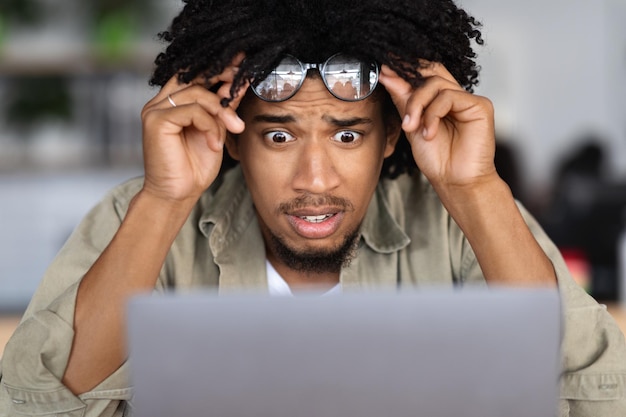 Il maschio riccio millenario afroamericano scioccato si toglie gli occhiali e guarda il laptop nella caffetteria