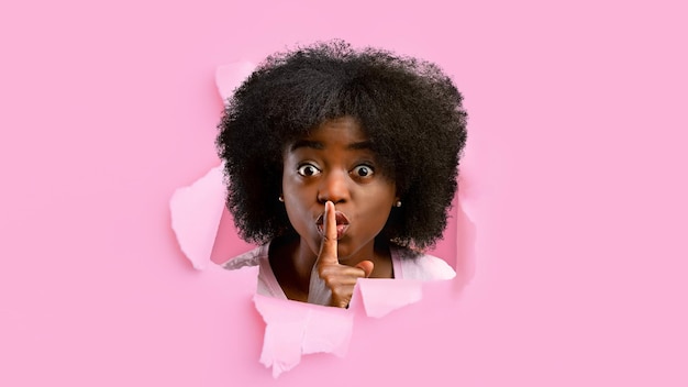 Shocked millennial black female looks into hole in pink paper shows finger near lips sign of silence