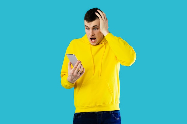 Shocked millennial guy holding smartphone reading negative message blue background