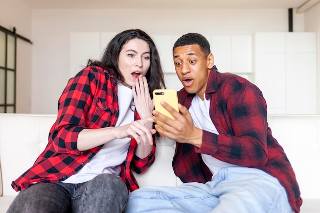 Shocked multiracial couple sitting on sofa at home and looking at smartphone