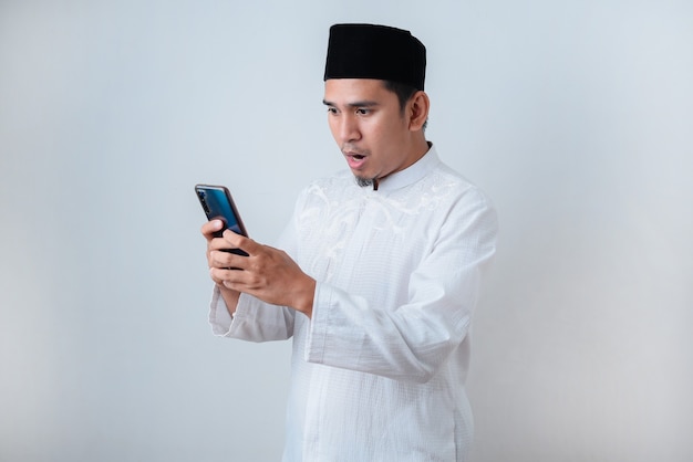 Shocked Muslim man wearing muslim clothes holding mobile phone look at phone screen against on white wall