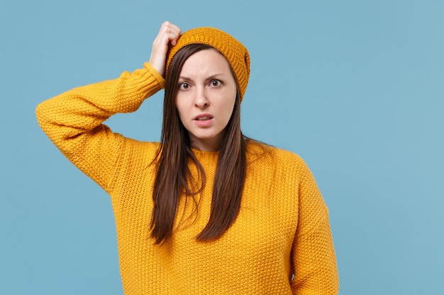 Scioccato perplesso giovane donna bruna ragazza in maglione giallo e cappello in posa isolato su sfondo blu ritratto in studio. concetto di stile di vita delle emozioni delle persone. mock up spazio di copia. mettere la mano sulla testa.