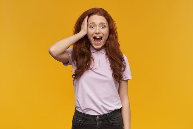 Shocked, positive woman with long ginger hair. wearing pink t-shirt. people and emotion concept. touching her head, surprised. isolated over orange wall