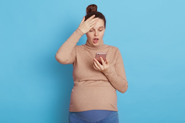 Foto donna incinta scioccata alla ricerca di uno schermo dello smartphone con la bocca aperta, toccando la sua testa isolata sopra la parete blu.