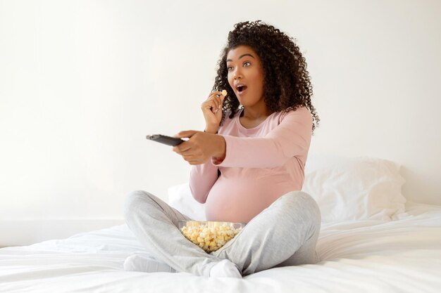 Шокированная беременная женщина с попкорном и пультом дистанционного управления реагирует на контент по телевизору