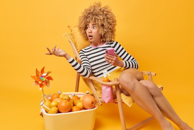 Shocked puzzled woman looks confused away shrugs shoulders\
wears casual clothes holds mobile phone chats in social networks\
poses on deck chair near portable fridge isolated over yellow\
background