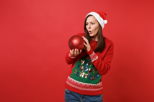 Shocked puzzled young Santa girl looking on big shiny tree toy in hands, keeping mouth wide open isolated on red background. Happy New Year 2019 celebration holiday party concept. Mock up copy space.