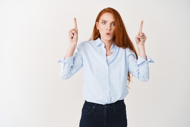 Shocked redhead woman showing something disturbing, pointing fingers up and staring confused at front, standing against white wall