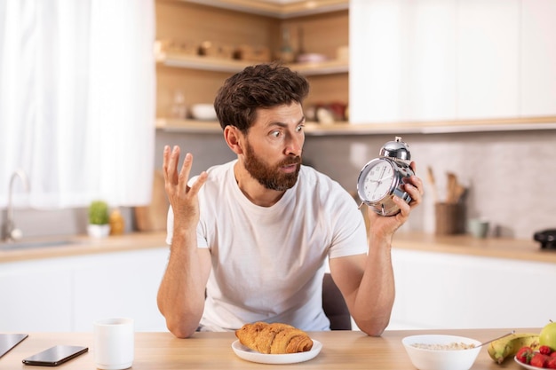 Shocked sad adult caucasian man with beard in white tshirt\
looks at alarm clock and late to meet or work