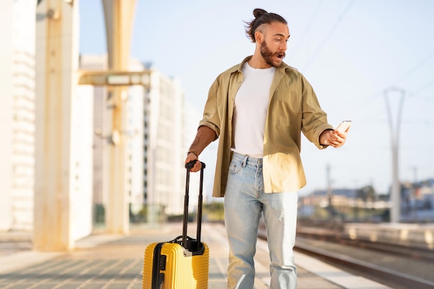 スーツケースを持ったショックを受けた悲しいミレニアル世代の白人男性が駅で電話を見る