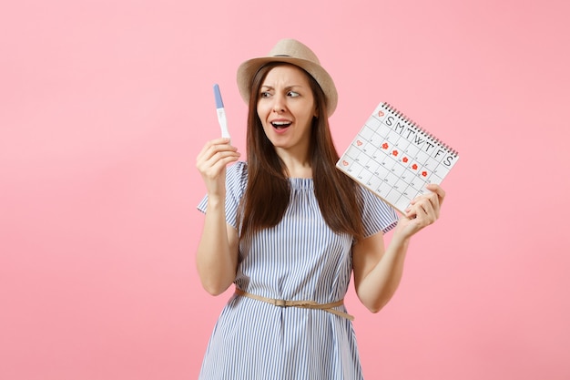 Shocked sad woman in blue dress, hat hold in hand pregnancy test, periods calendar for checking menstruation days isolated on pink background. Medical, healthcare, gynecological concept. Copy space.