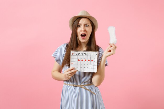Shocked sad woman in blue dress, hat holding sanitary napkin, female periods calendar for checking menstruation days isolated on pink background. Medical, healthcare, gynecological concept. Copy space