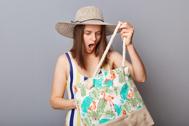 縞模様のワンピース水着と日焼け止めを探している彼女のバッグを見て灰色の背景にポーズをとるわらの広いつばの帽子をかぶったショックを受けた怖い女性