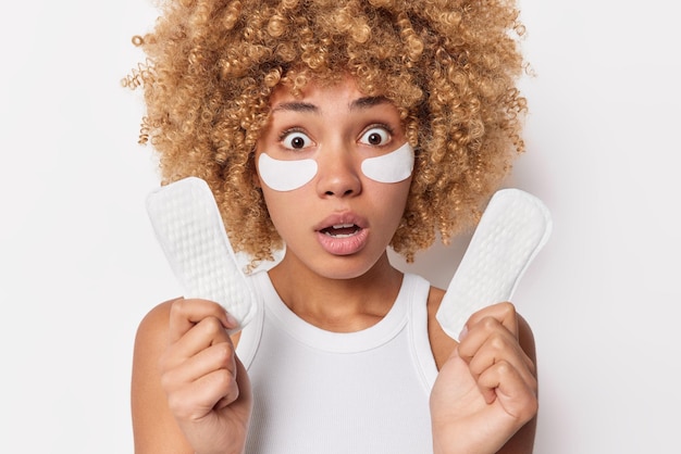 Shocked scared woman with blonde curly hair stares bugged eyes holds sanitary napkins applies beauty patches under eyes for skin treatment dressed in casual t shirt isolated over white wall