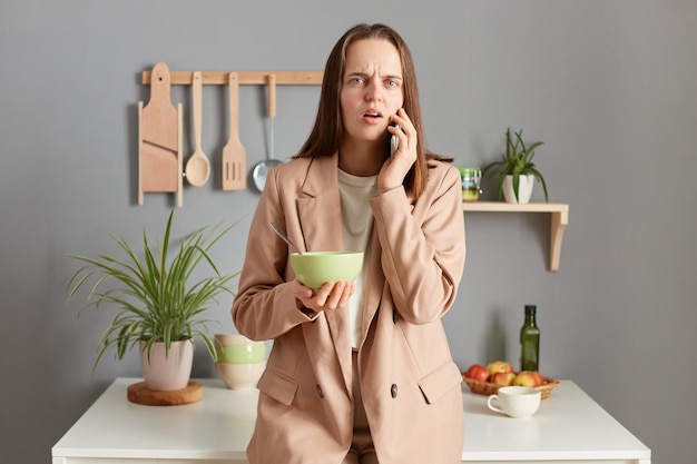 Shocked scared woman with brown hair wearing beige jacket standing in home kitchen interior talking on smart phone while having breakfast hearing bad news