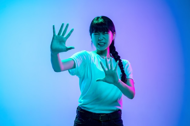 Shocked, scared. Young asian woman's portrait on gradient blue-purple studio background in neon light. Concept of youth, human emotions, facial expression, sales, ad. Beautiful brunette model.