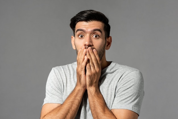 Shocked scared young caucasian man with eyes popping and hands covering mouth on light gray studio background