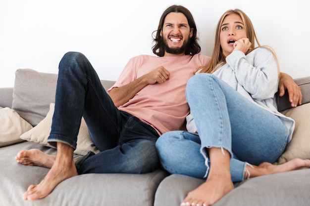 a shocked scared young loving couple on sofa watch tv film indoors at home.