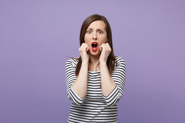 Shocked scared young woman in casual striped clothes posing isolated on violet purple background in studio. People lifestyle concept. Mock up copy space. Keeping mouth open, clenching fists near face.