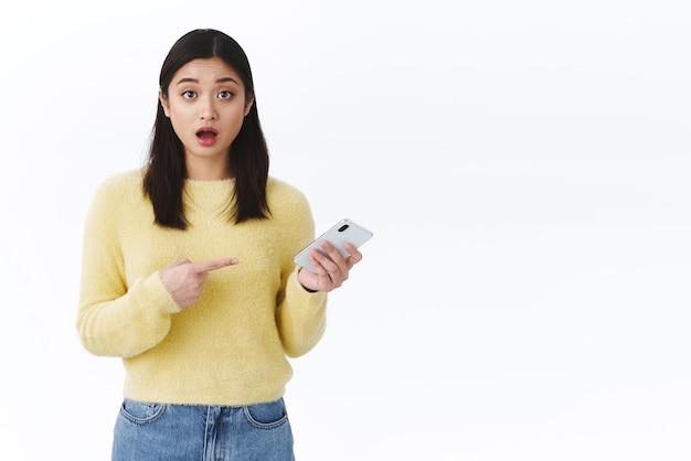 ショックを受けた言葉のないアジアの女の子が口を開けて携帯電話を指差して、新しいスマートフォン機能の白い背景で助けを求める最後のニュースについてオンラインで話している驚くべき何かを見て
