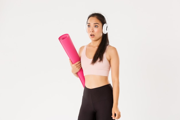 Shocked and startled asian girl in headphones, holding rubber mat, stop and drop jaw.