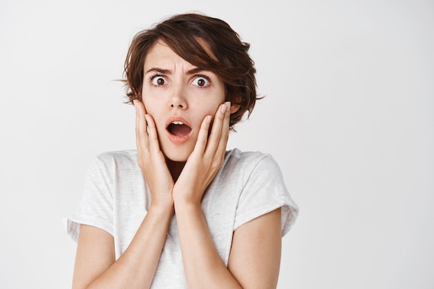 Shocked and startled caucasian woman gasping and frowning, looking at something terrible, standing on white wall in casual t-shirt