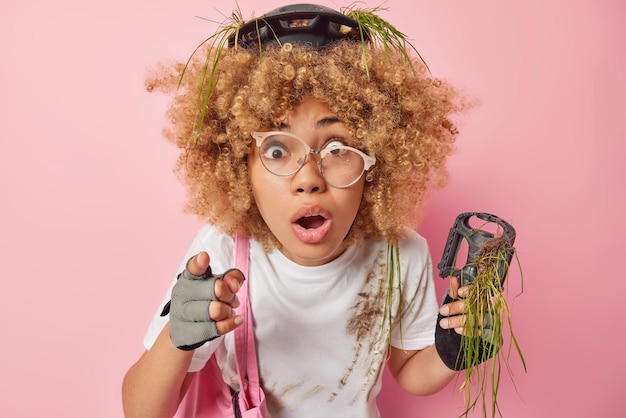 Shocked startled curly woman wears helmet gloves and dirty t\
shirt points directly at camera cannot believe own eyes got into\
bicycle accident poses against pink background omg just look\
there