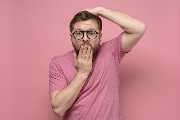 Shocked strange man with glasses in fright covers mouth with one hand put the other on head