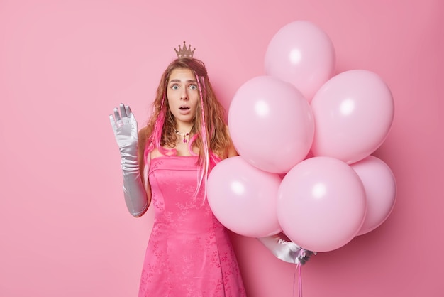 Shocked stunned young woman wears festive dress and gloves\
holds big bunch of helium balloons comes on birthday party reacts\
on something amazing isolated over pink background holiday\
concept