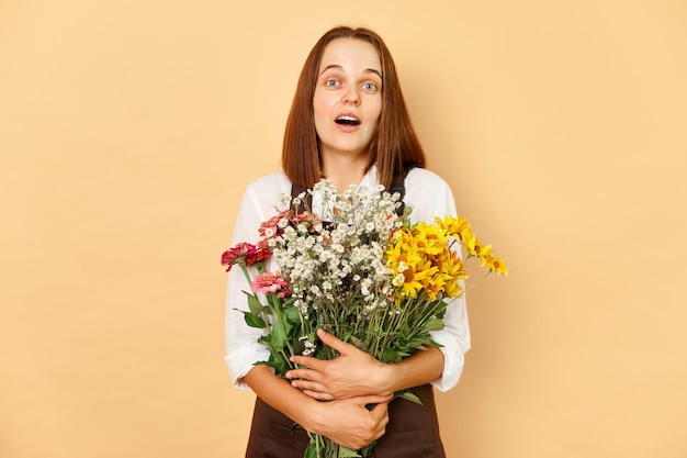 Photo shocked surprised amazed adult woman florist wearing brown apron holding bouquet standing isolated over beige background with beautiful flower composition