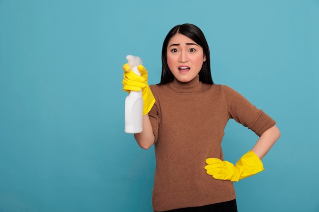 Shocked surprised asian housekeeper from chores holding spray\
in yellow glove ready for duty, cleaning home concept, amazed\
wondering woman and negative from day to day work