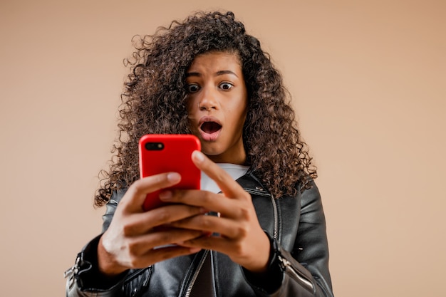 Shocked surprised black girl looking at her phone screen isolated over brown