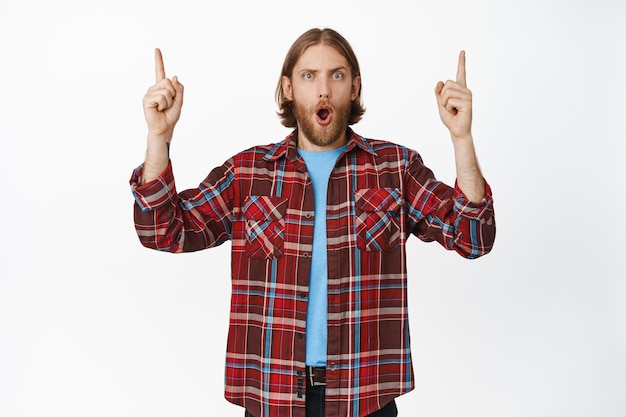 Shocked and surprised blond adult man drop jaw, gasping WOW face, pointing fingers up at top sale, advertisement, showing big promo, standing impressed against white background