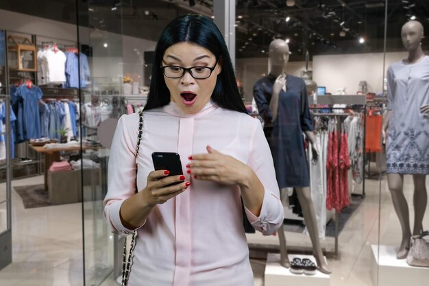 Shocked surprised brunette asian woman looking at her smartphone clothing boutique shop in the backg