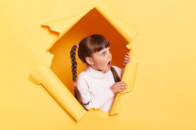 Shocked surprised little girl with braids wearing casual shirt looking through torn hole in yellow paper looking aside with open mouth sees shocking advertisement