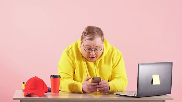 Shocked and surprised man in yellow sportswear is sitting at a table watching the news on a pink background on his smartphone