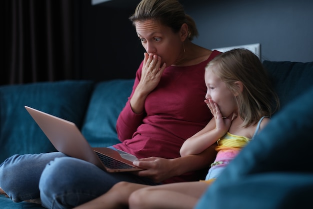 Шокированные удивленные мама и дочь, глядя на экран ноутбука, сидя на диване
