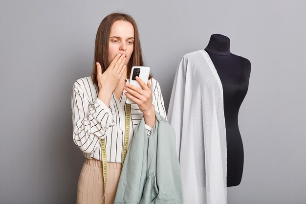 Shocked surprised scared professional female seamstress with mobile phone in hands covering mouth with hand wrong order dressmaking mistake