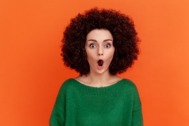 Shocked surprised woman with Afro hairstyle wearing green casual style sweater looking at camera with big eyes and open mouth, sees astonishing things. Indoor studio shot isolated on orange background