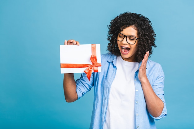 Shocked surprised young curly african american woman isolated on blue background studio portrait. Mock up copy space. Holding gift certificate.