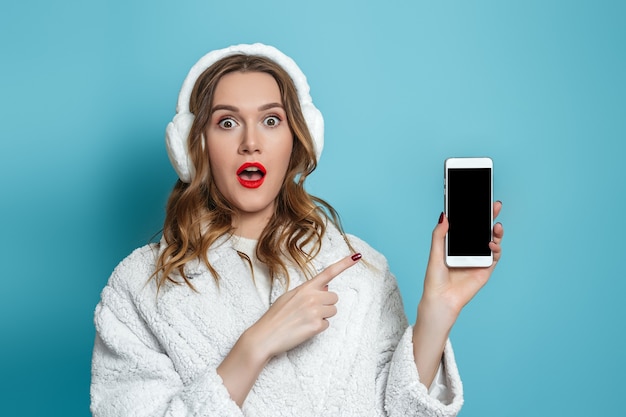 흰색 인조 모피 코트 포인트 손가락 블루 스튜디오 배경에 고립 된 휴대 전화 화면에서 충격 된 놀된 젊은 여자.