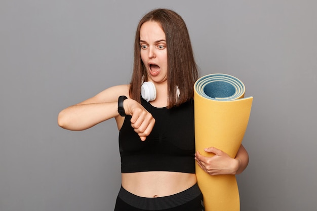 Shocked surpruised woman holding rolled fitness mat dressed in activewear isolated over gray background looking at smartwatch with widely opened mouth checking burning calories