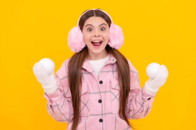 Shocked teen girl in fur earmuffs playing snowballs on yellow background winter