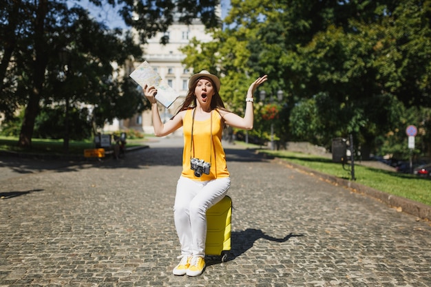 노란색 옷을 입은 충격적인 여행자 관광 여성, 여행 가방에 앉아 있는 모자는 도시 야외에서 손을 펼치고 있는 도시 지도를 들고 있습니다. 주말 휴가를 여행하기 위해 해외로 여행하는 소녀. 관광 여행 라이프 스타일.