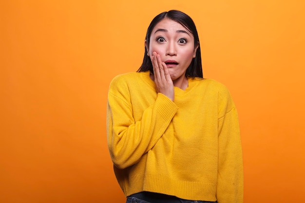 Shocked troubled asian woman wearing yellow vibrant sweater while putting hand on face. Uneasy young adult girl with shock expression, wearing vivid trendy clothing while on orange background.