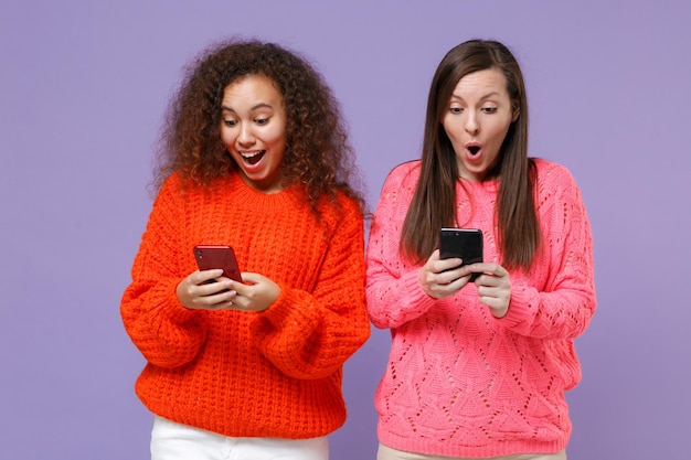 Shocked two young european african american women friends in
knitted sweaters isolated on violet purple background. people
sincere emotions lifestyle concept. using mobile phone, typing sms
message.