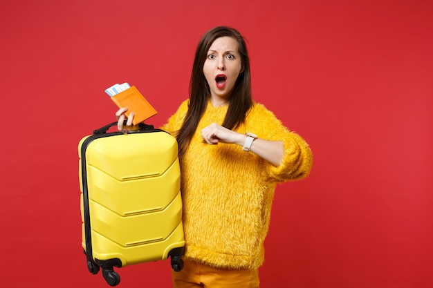 Shocked upset woman in yellow fur sweater with opened mouth late hold suitcase passport boarding pass ticket isolated on red background. People sincere emotions, lifestyle concept. Mock up copy space.