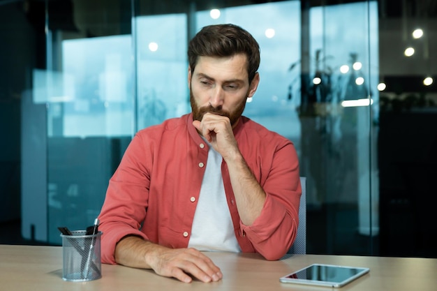 Photo shocked and upset young man sitting at the desk in the office thoughtfully holding his head with his